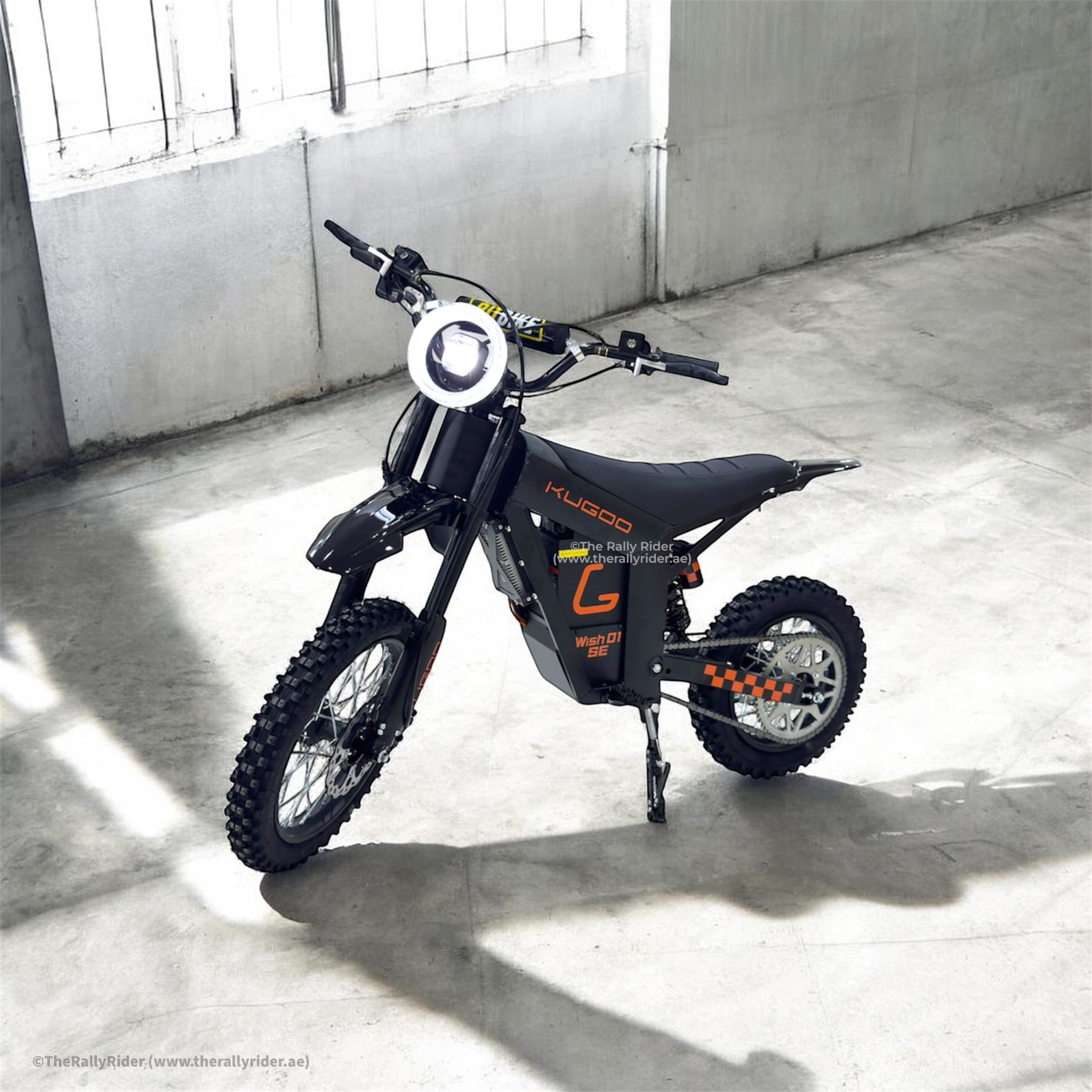 Kugoo Wish 01 SE electric dirt bike parked on a concrete floor indoors, round LED headlight glowing, black body with orange graphics and knobby tires.